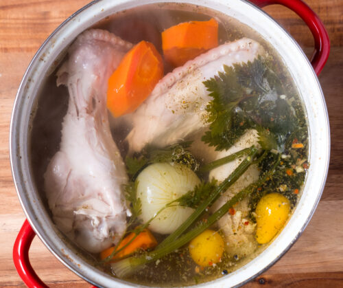 Turkey broth with pieces of meat on bone and vegetables in cooking pot on a wooden table, view from